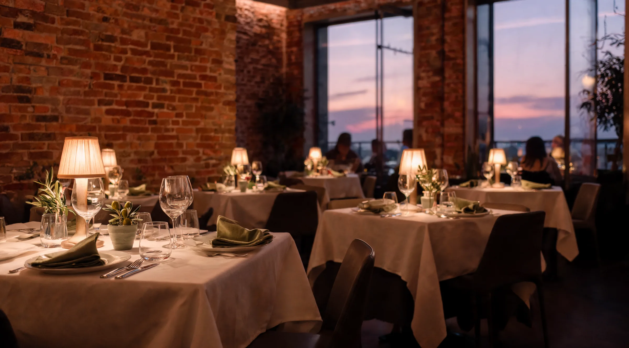 Candlelit dining room at Locanda, tables set with linen and olive branches, sunset through the windows